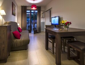 A modern living room with a sofa, a dining table, a TV, and a view through a glass door. - Stay in Les Eyzies With Pool (Les Eyzies)