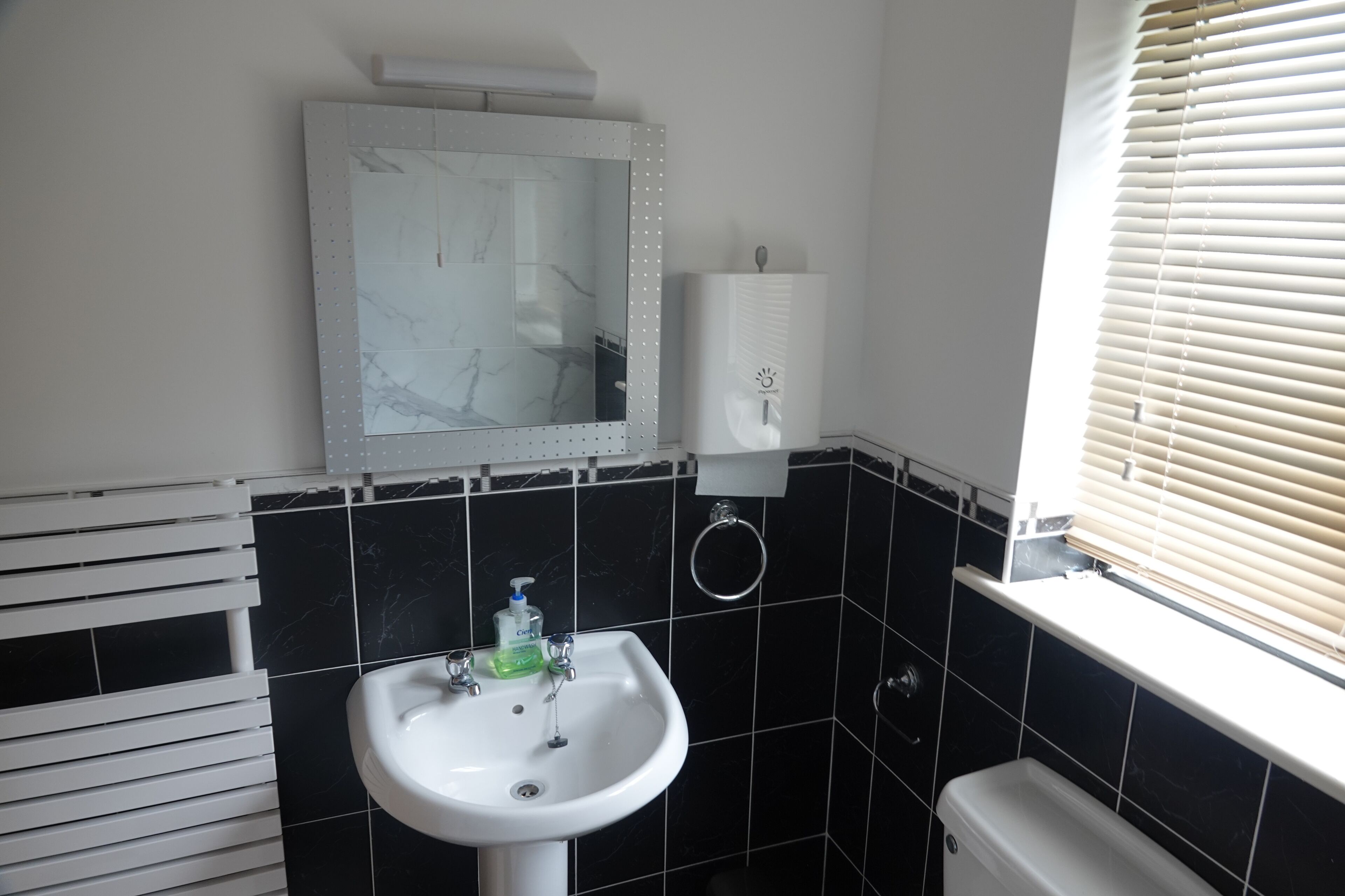 Ein Badezimmer mit weißem Waschbecken, einem Spiegel, einem Handtuchhalter, einer Toilette und einem Fenster mit Jalousien.