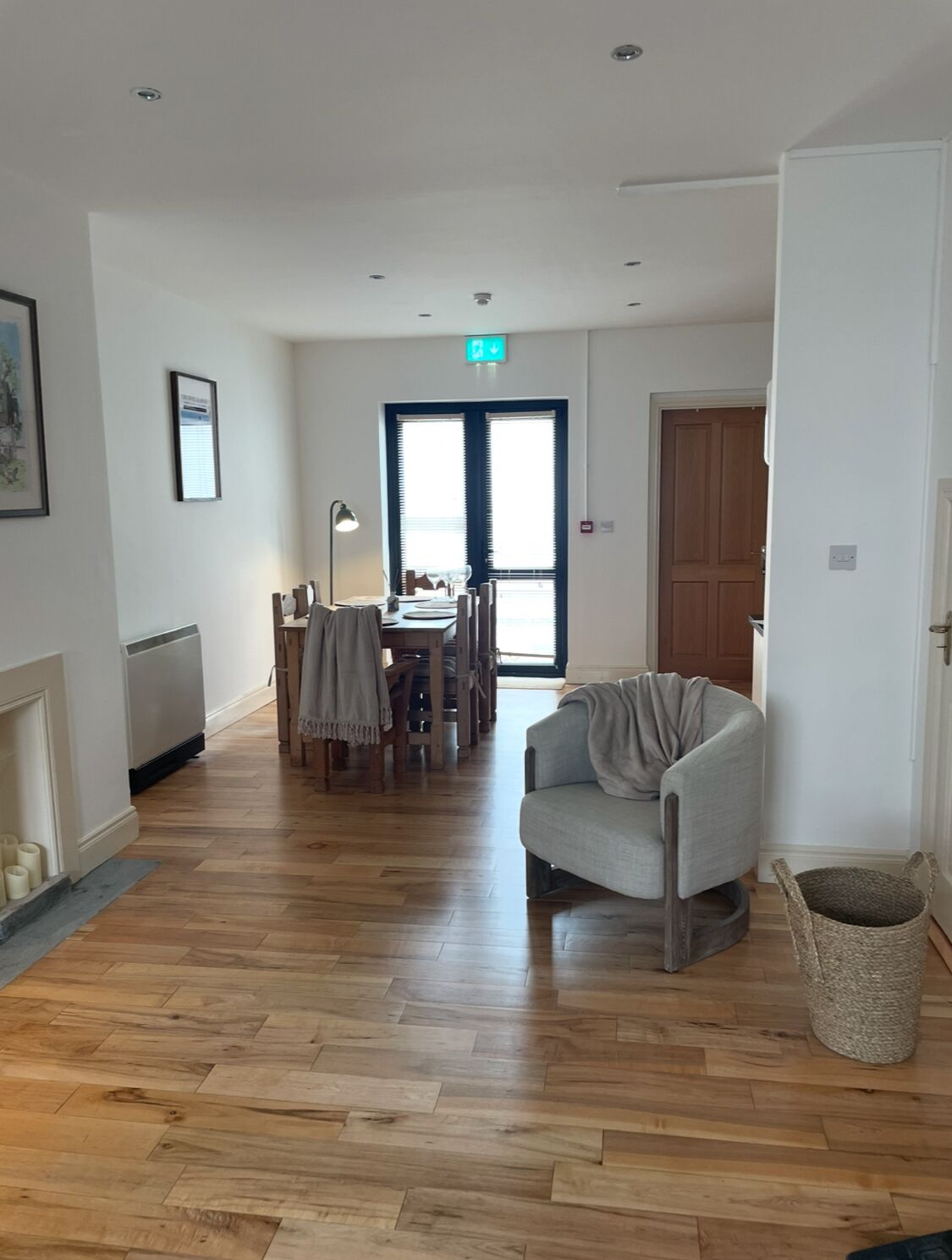 A room with a wooden floor, a grey armchair, a dining table with chairs, a fireplace, and a large window with blinds.