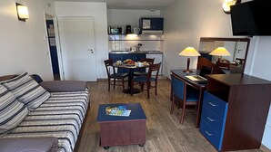 Ein kleines Hotelzimmer mit Bett, Schreibtisch, Stuhl, einer kleinen Küchenzeile und einem Fernseher.