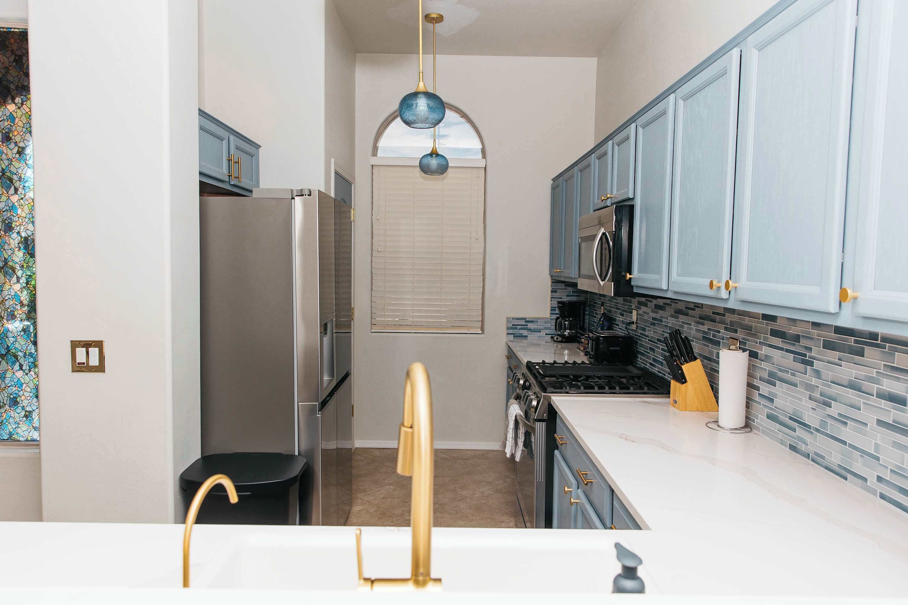 A kitchen with a refrigerator, stove, and microwave. There is a window with blinds, a sink with a gold faucet, and cabinets with gold handles.