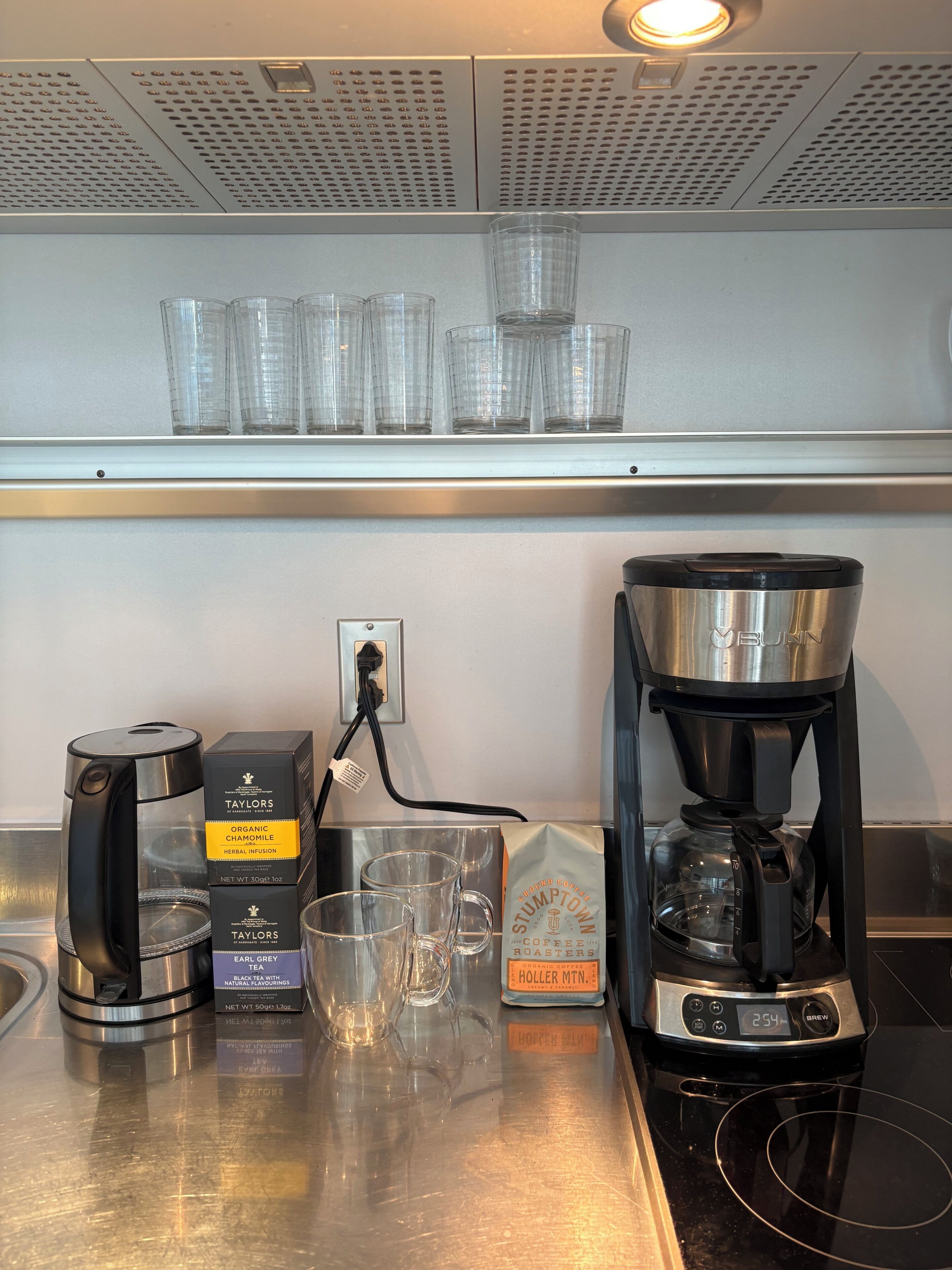 Een koffiehoek met een koffiezetapparaat, theemaker en glazen bekers op een roestvrijstalen toonbank.