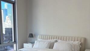 A modern bedroom with a large bed, a nightstand, a lamp, and a view of the city through a large window. - Rockefeller Center 1 Bedroom with in unit W&D, Gym & amazing views (New York)