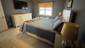 Ein Schlafzimmer mit einem Bett, einem Deckenventilator, einem Fenster mit Jalousien, einem Schminktisch und einem an der Wand befestigten Fernseher.
