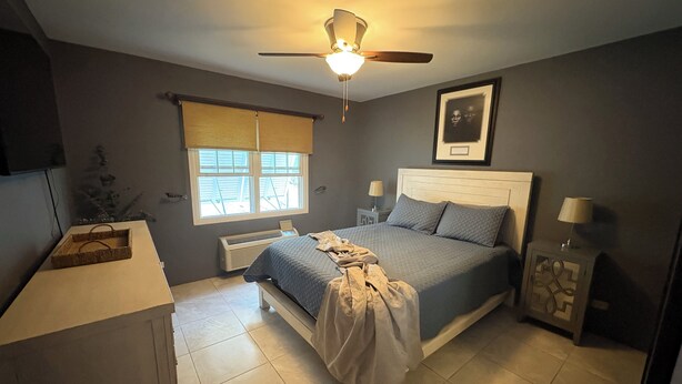 Ein Schlafzimmer mit Bett, Nachttischlampen, Deckenventilator, Fenster mit Jalousien, Heizkörper und einem gerahmten Bild an der Wand.