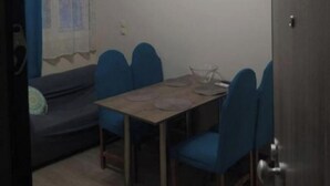 Quarto compacto com mesa de jantar, cadeiras azuis, sofá e janela com cortina.