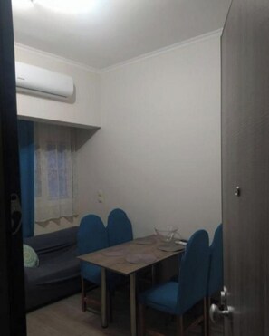 A compact room with a dining table, blue chairs, a sofa, and a window with a curtain. - 2-bedroom apartment in marvelous Loutraki with AC comfort (Loutraki)