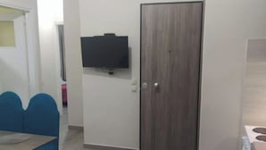 Quarto com porta de madeira, televisão de tela plana na parede, mesa de jantar com cadeiras azuis e área de cozinha com pia e bancada.