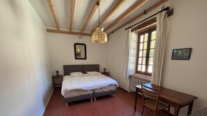 Een slaapkamer met een bed, nachtkastje, tafel, raam met gordijnen en een hanglamp.