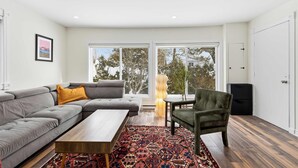 A modern living room with a grey sectional sofa, a wooden coffee table, a patterned rug, and a view of trees through large windows.