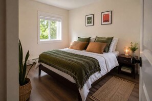 A bedroom with a bed, bedside tables, a nightstand, a lamp, a potted plant, and a window.