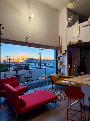 Ein moderner Wohnraum mit Balkon, roter Einrichtung, einem kleinen Tisch mit Obstschale und Blick auf einen Sonnenuntergang über einer Brücke.