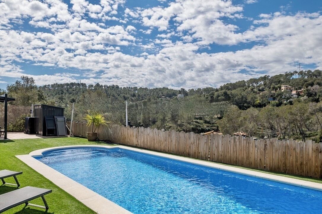 A swimming pool with a wooden fence, a grassy area, and a hill with trees in the background.