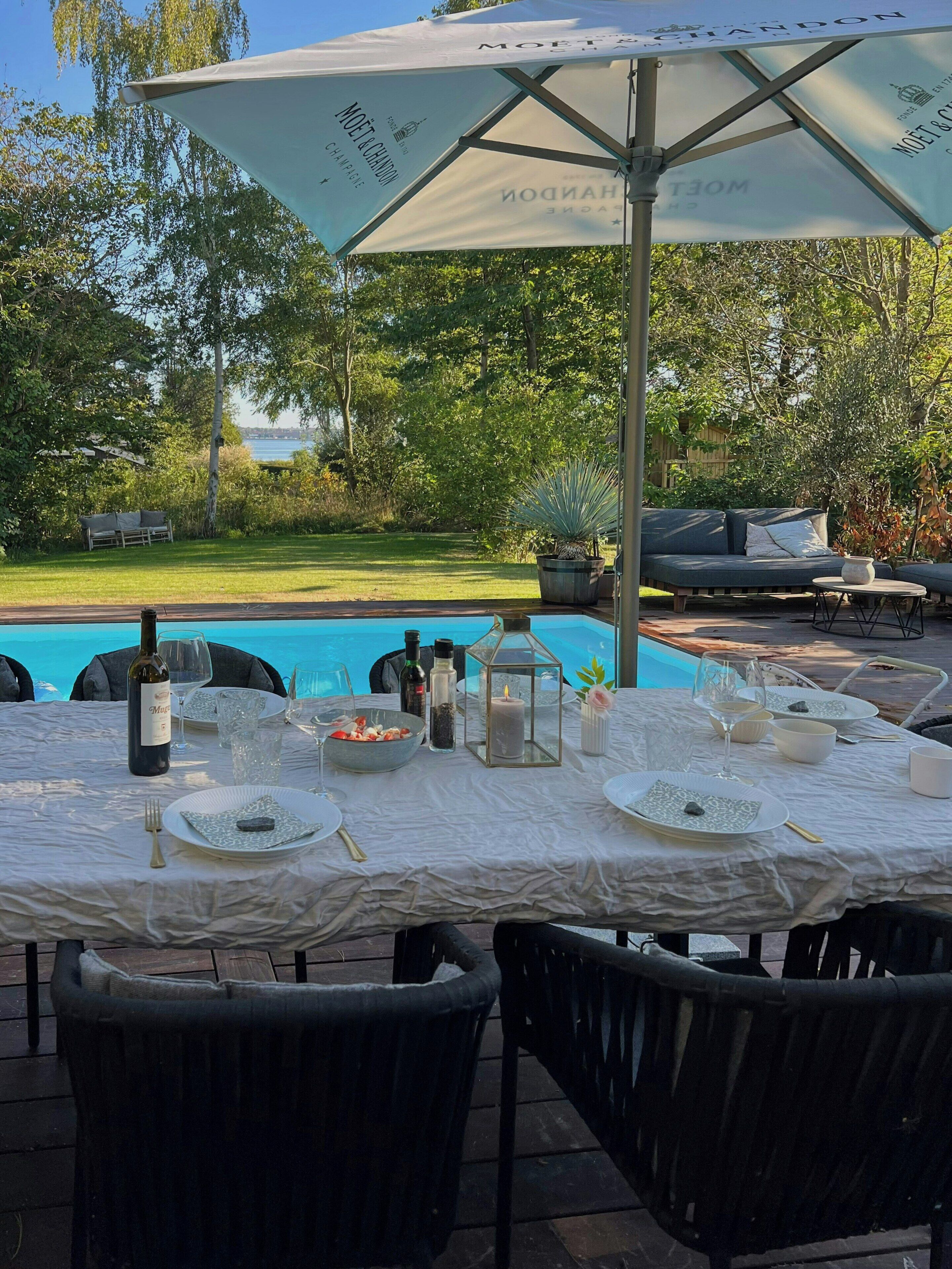 An outdoor dining area with a pool view, a table set with plates, glasses, and a bottle, surrounded by chairs.