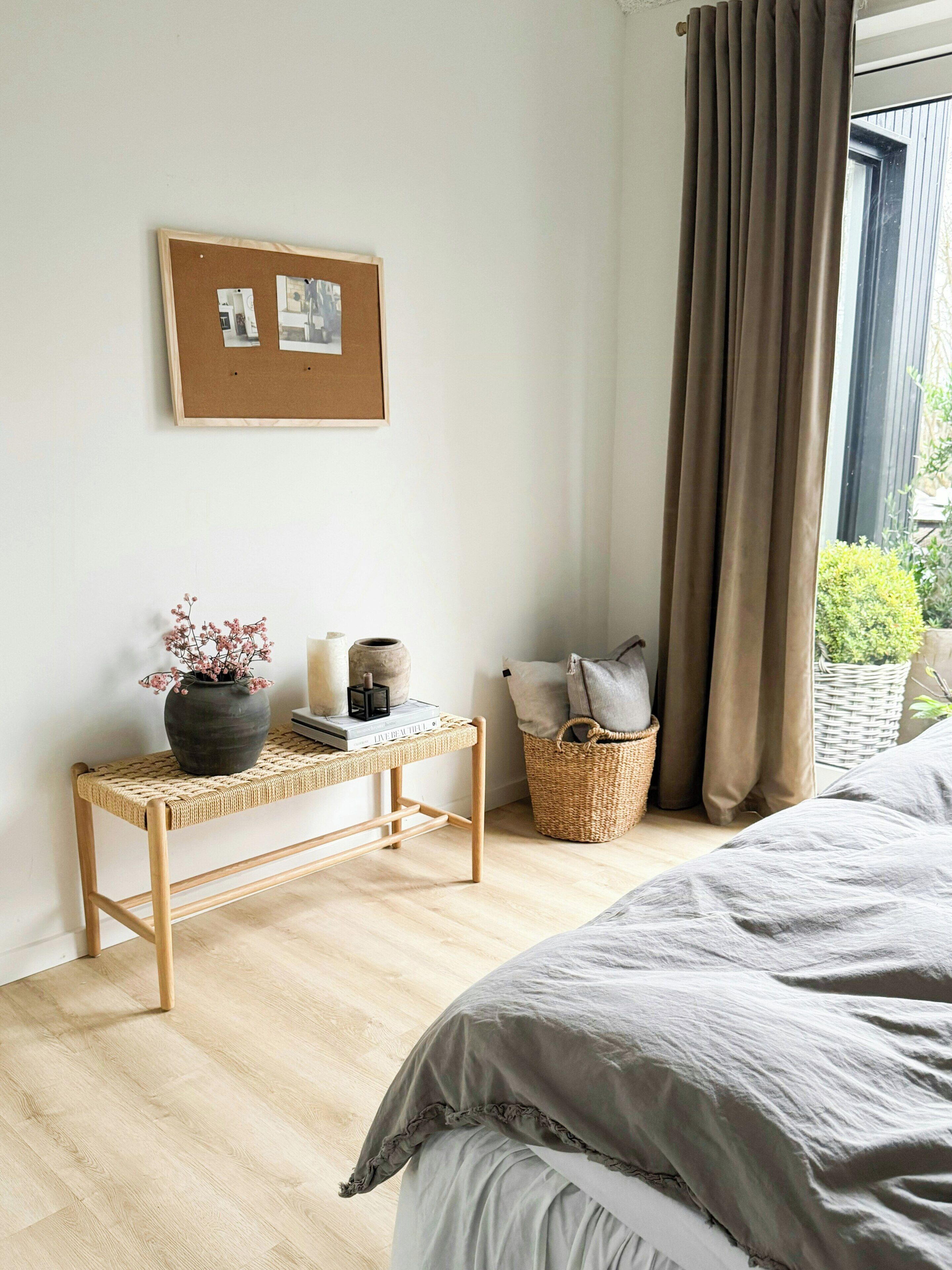 A bedroom with a bed, a bedside table with a vase and books, a framed picture on the wall, and a window with curtains.