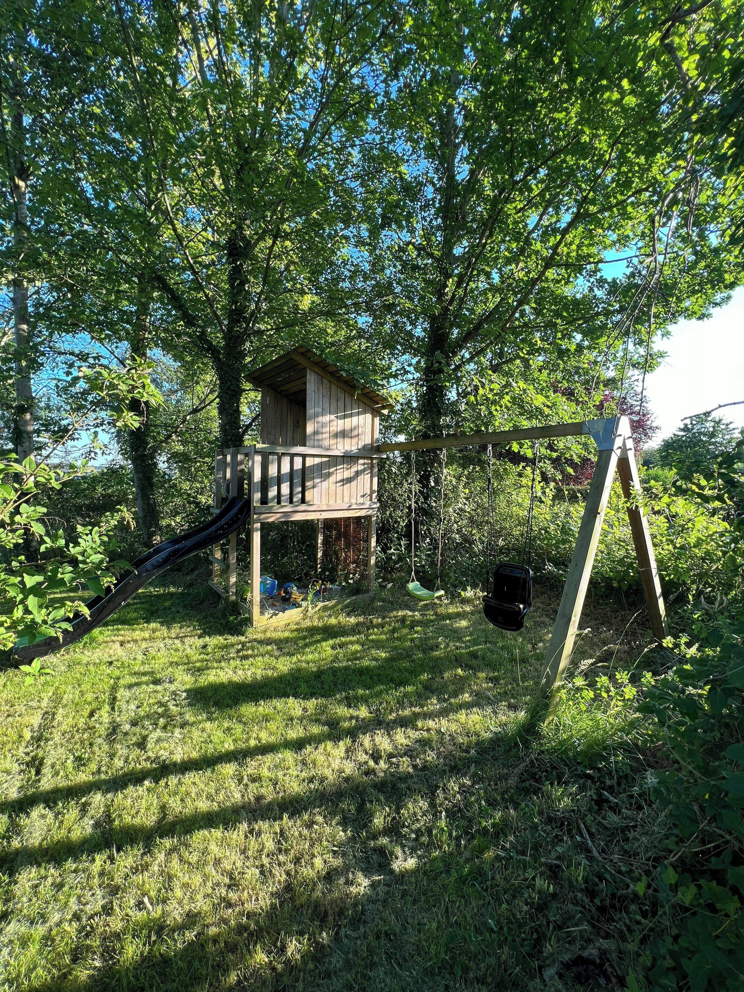 A wooden playhouse with a slide, a swing, and a swing set in a grassy area with trees.