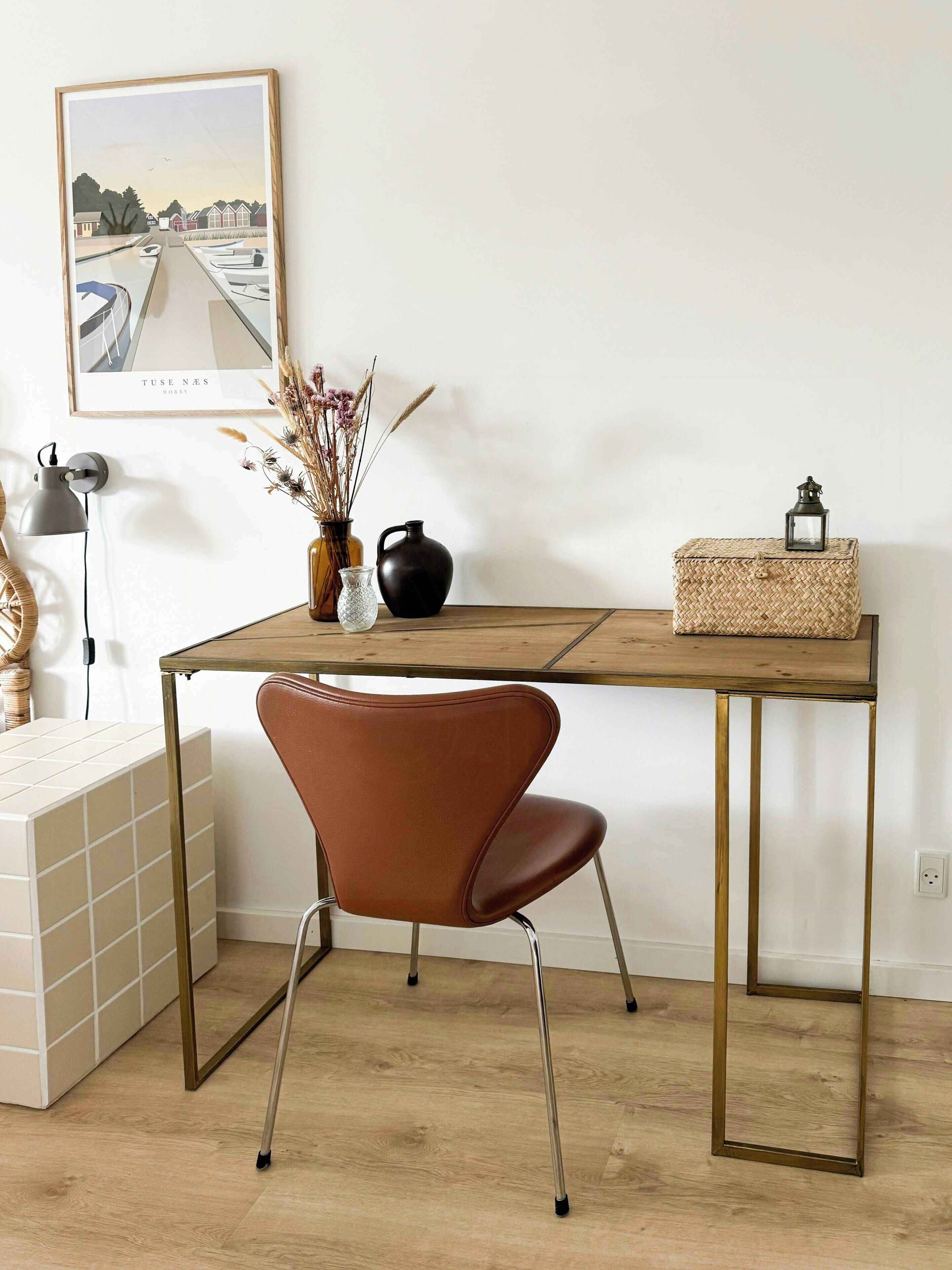 A wooden desk with a chair, a framed picture, a vase with dried flowers, a black pitcher, and a woven basket.
