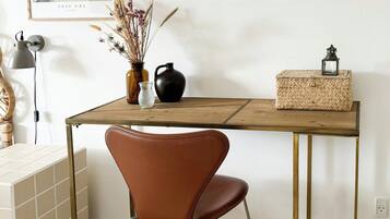 A wooden desk with a chair, a framed picture, a vase with dried flowers, a black pitcher, and a woven basket.
