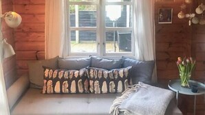 Et hyggeligt interiør i en hytte med træloft, et vindue med gardiner, en sofa med mønstrede puder, et lille bord med en vase og en lampe.