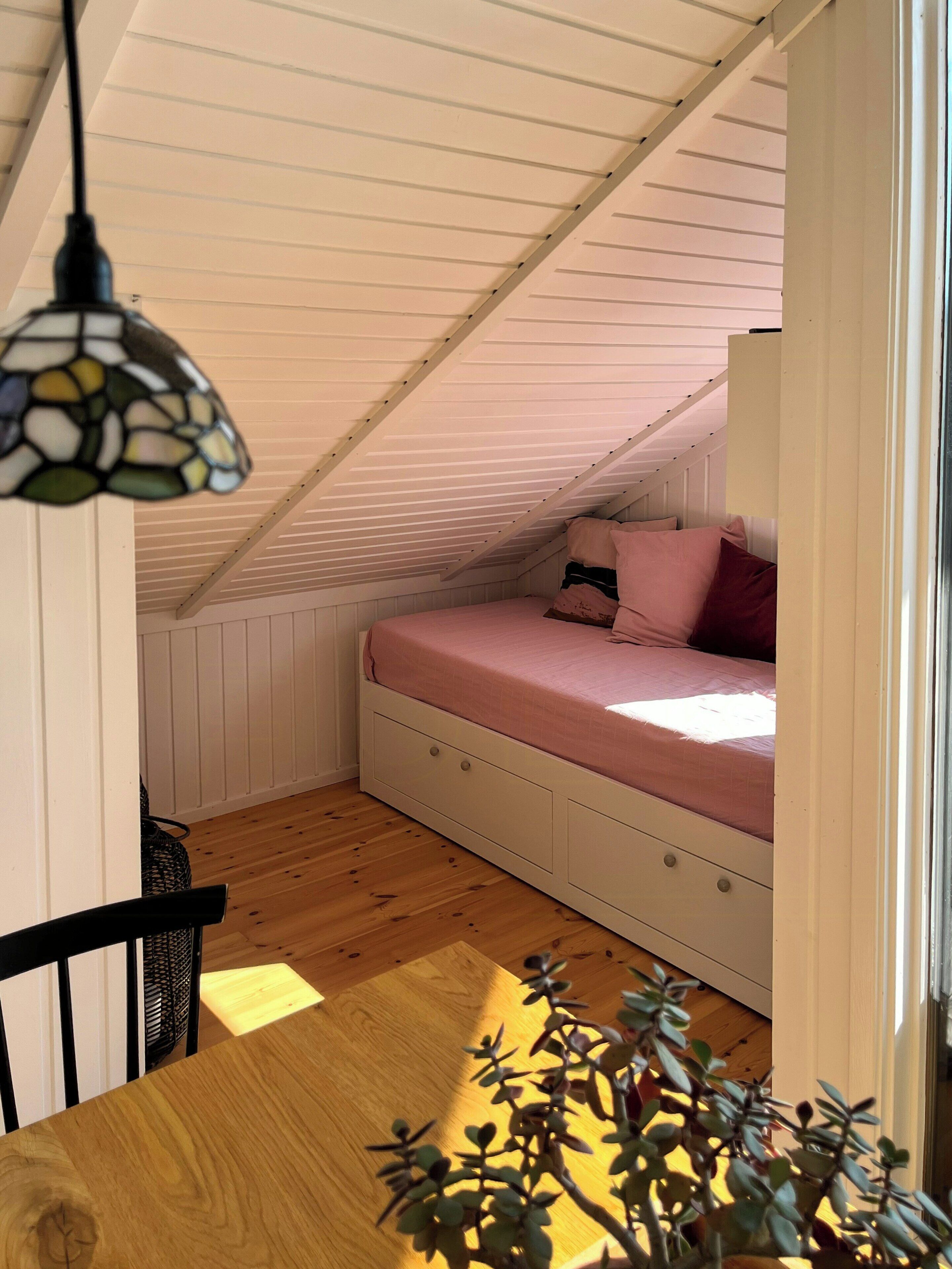 A small attic bedroom with a sloped ceiling, a bed with pink bedding, a wooden table, and a potted plant.