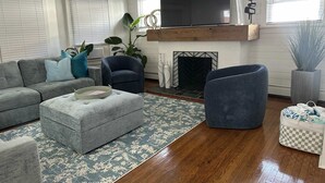 A living room with a sectional sofa, a blue armchair, a small ottoman, a fireplace, and a television.