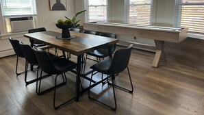 A dining area with a wooden table, black chairs, a vase with green plants, and large windows with blinds.