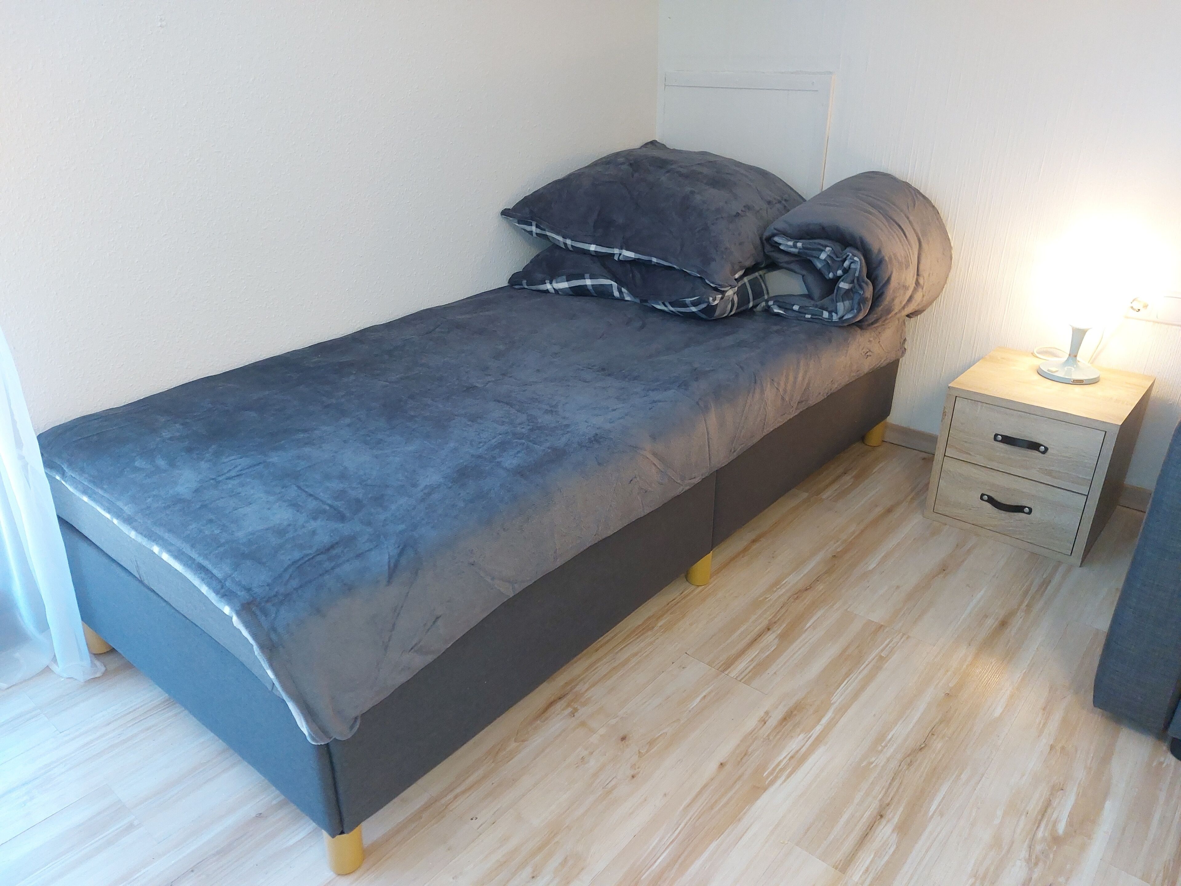 Una cama individual con colchón y almohadas grises, una mesita de noche con lámpara y suelo de madera.