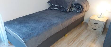 Una cama individual con colchón y almohadas grises, una mesita de noche con lámpara y suelo de madera.