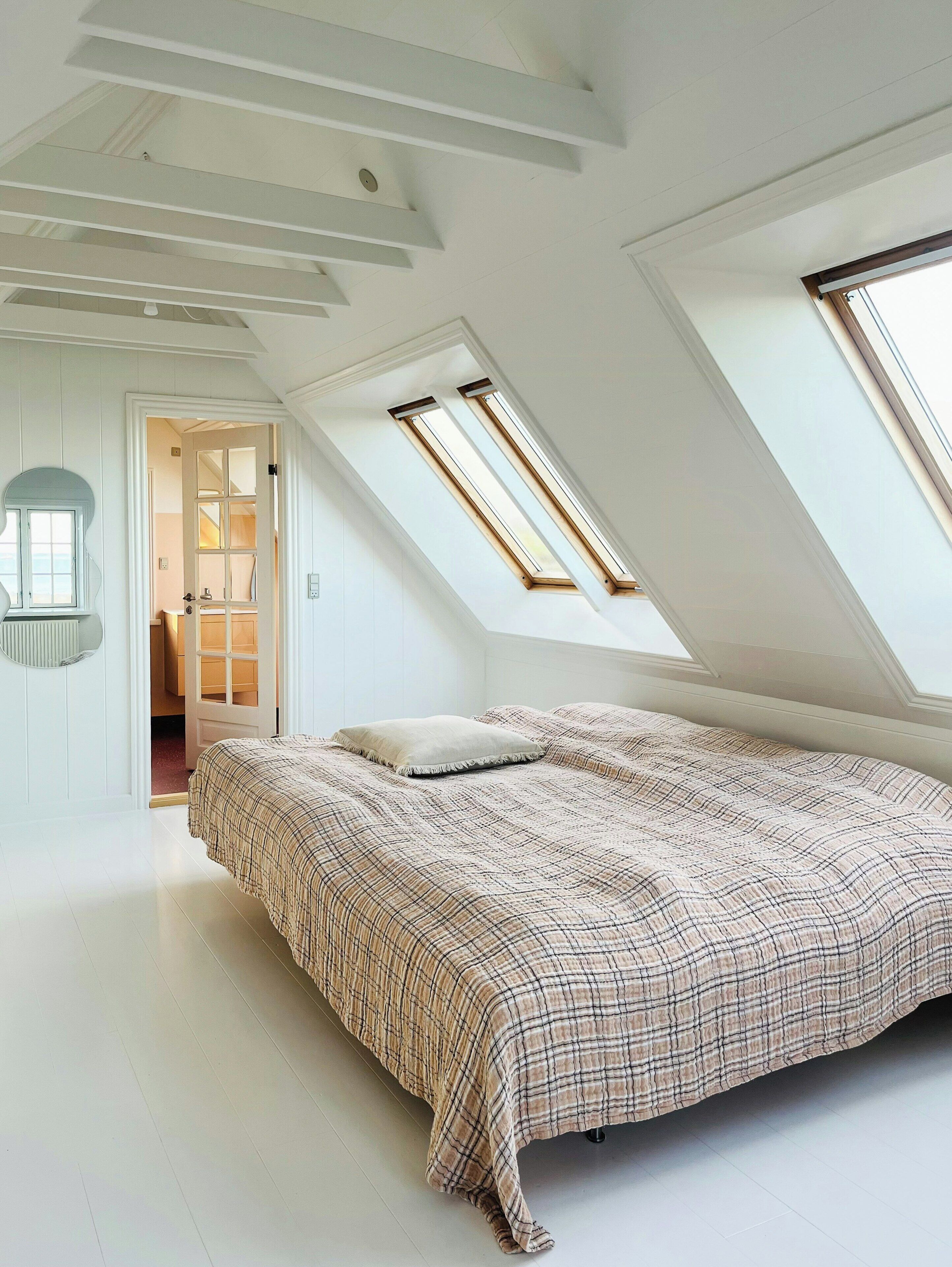 A bedroom with a sloped ceiling, skylights, a bed with a patterned bedspread, and a door leading to another room.
