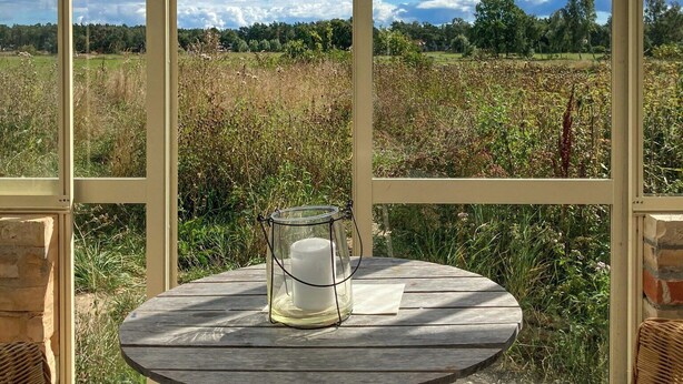 A room with a large window, a round table with a candle holder, wicker furniture, and a view of a field. - Holiday home with greenhouse near Mälarhusen (Skåne län)