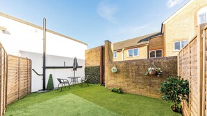 A backyard with a lawn, a table, chairs, a hanging plant, and a brick wall. - Two bed High Street Apartment (Teddington)