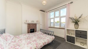 A bedroom with a bed, a fireplace, a window with curtains, and a bedside table with drawers. - Two bed High Street Apartment (Teddington)