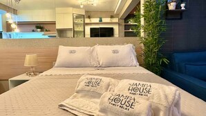 A modern hotel room with a large bed, a nightstand, a lamp, a TV, and a view of the kitchen and living area. - Jampa House - Your beachfront apartment in Bessa (João Pessoa)