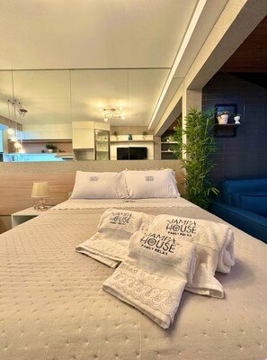 A modern hotel room with a large bed, a nightstand, a lamp, a TV, and a view of the kitchen and living area. - Jampa House - Your beachfront apartment in Bessa (João Pessoa)