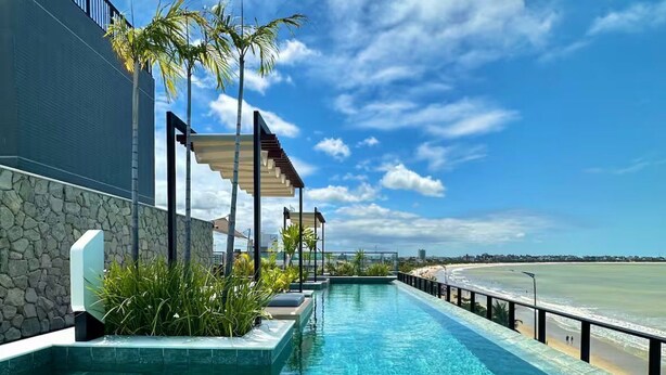 A swimming pool with clear water, a stone wall, and a view of the beach. - Jampa House - Your beachfront apartment in Bessa (João Pessoa)