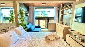 A hotel room with a bed, a TV, a balcony with plants, and a view of buildings. - Jampa House - Your beachfront apartment in Bessa (João Pessoa)