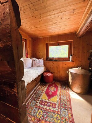 Ein gemütliches Cabin-Zimmer mit einem Bett, einem Fenster, einem roten Hocker und einem großen Kupfertopf.