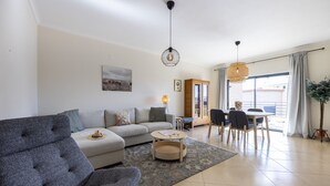 A modern living room with a grey sofa, a coffee table, and a dining area with chairs and a table. - Sea Breeze Lagos, Modern Apartment with Pool and Garden View – Lagos (Lagos)