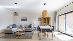 A modern living room with a sofa, coffee table, and dining table. There is a large window providing a view of the outside. - Sea Breeze Lagos, Modern Apartment with Pool and Garden View – Lagos (Lagos)
