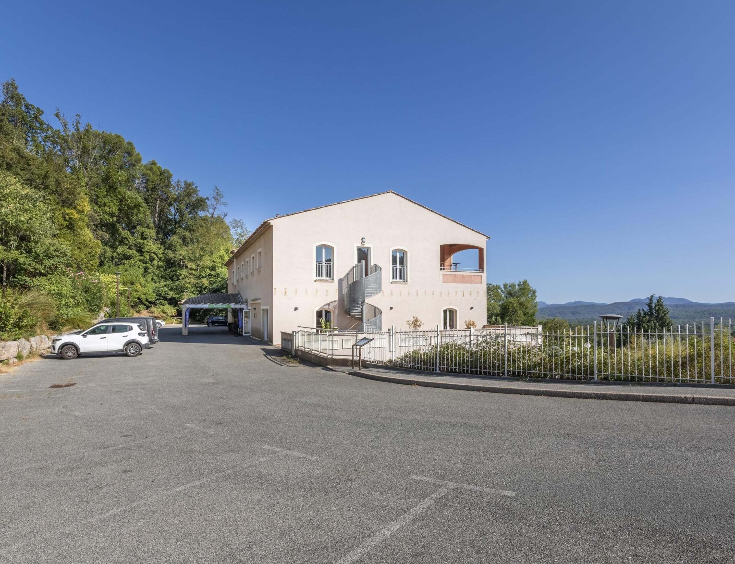 Un immeuble de deux étages doté d’un balcon, d’un escalier et d’un espace de stationnement où sont garées des voitures.