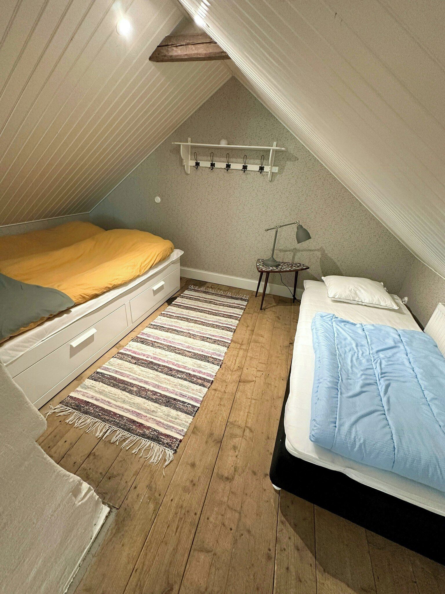 A small attic bedroom with two beds, a wooden floor, a striped rug, and a ceiling light fixture.