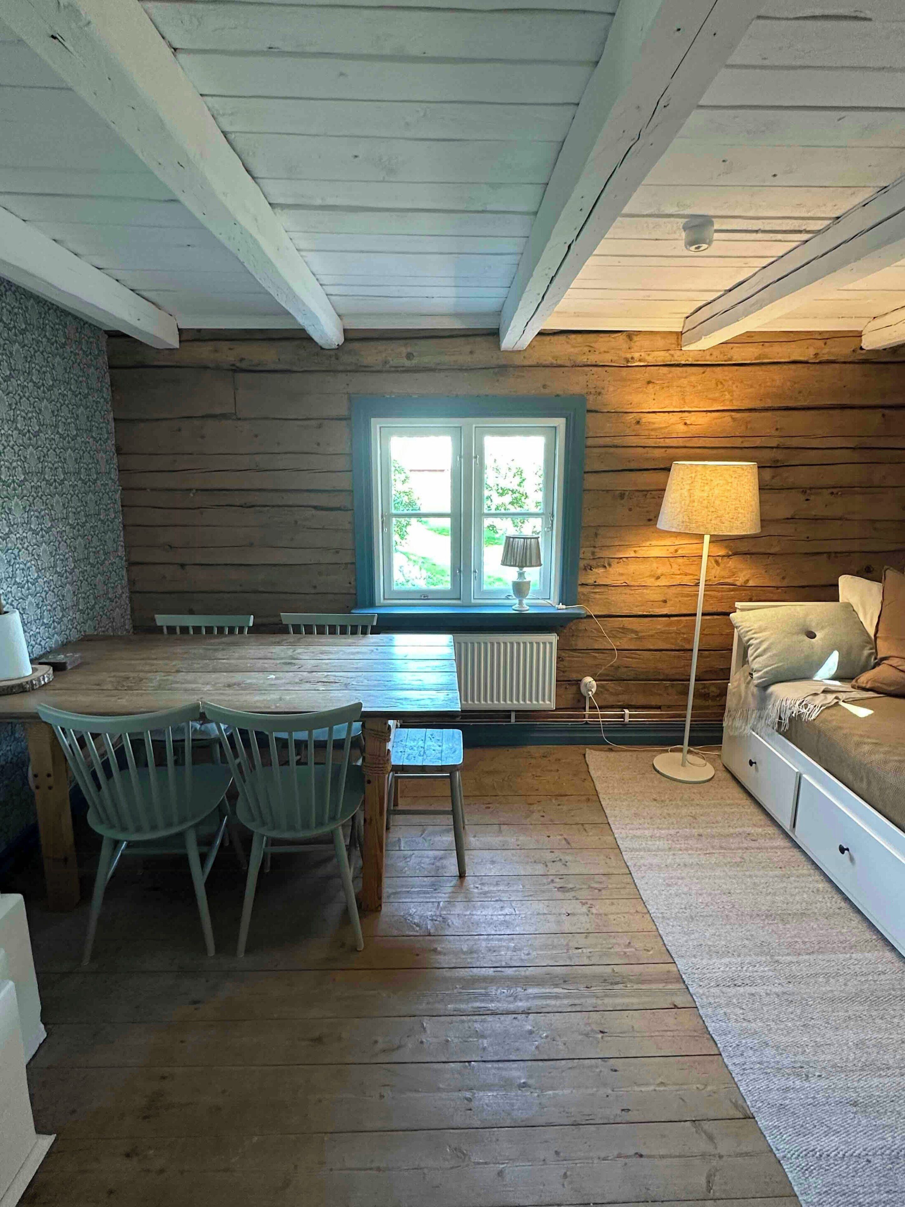 L’intérieur chaleureux d’un chalet avec une table à manger en bois, des chaises, une fenêtre donnant sur un paysage agréable, une lampe sur pied et un lit doté de tiroirs de rangement.