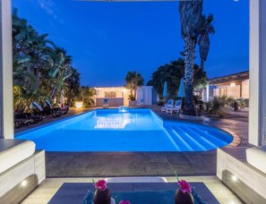 Ein Pool mit Liegestühlen und Palmen, umgeben von einer gut beleuchteten Terrasse.