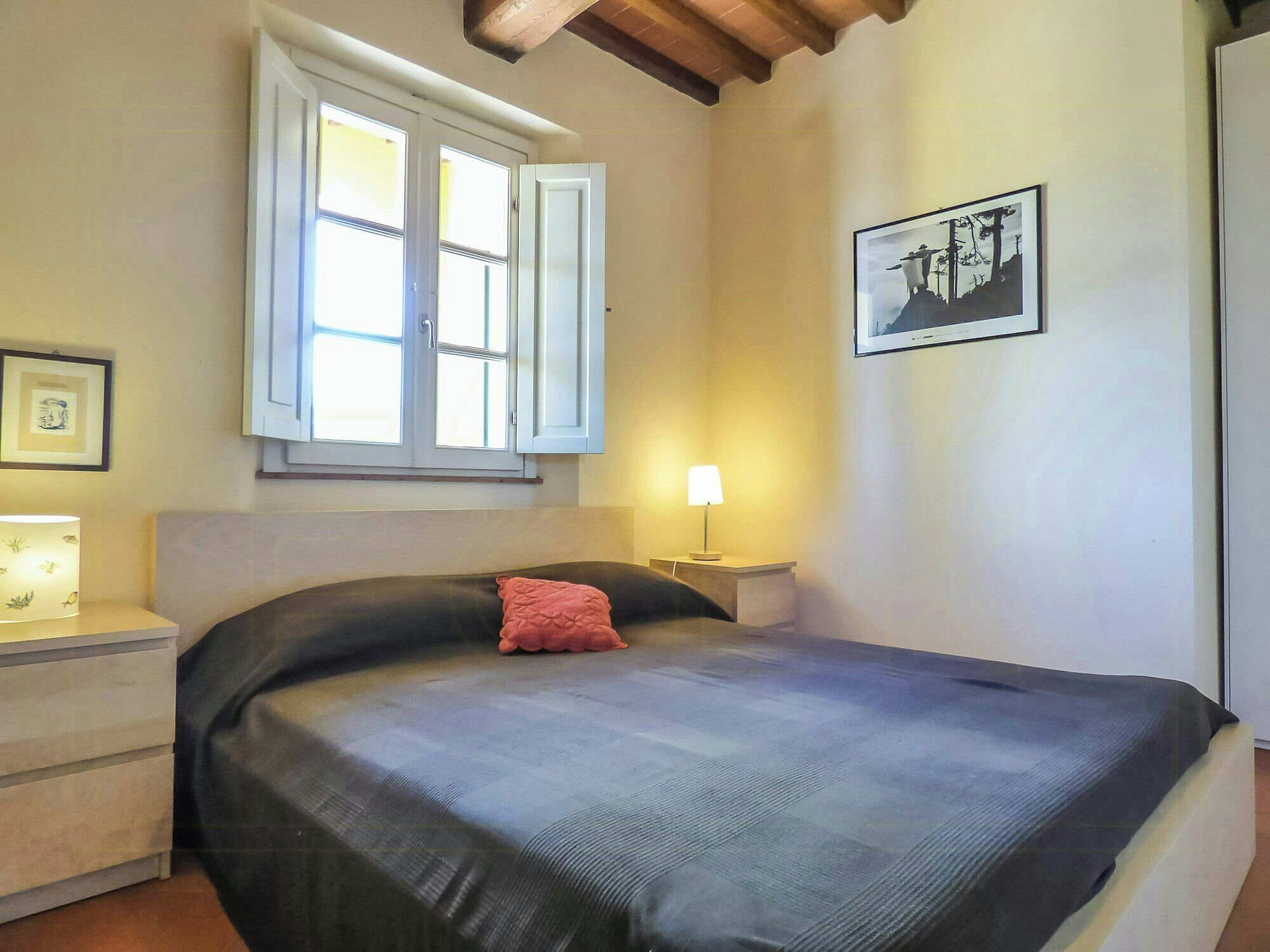 A bedroom with a bed, a window with shutters, a bedside lamp, a framed picture on the wall, and a nightstand.