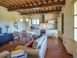 Eine moderne Küche mit weißen Schränken, eine Essecke mit Tisch und Stühlen sowie ein Wohnzimmer mit einer Couch und Büchern.