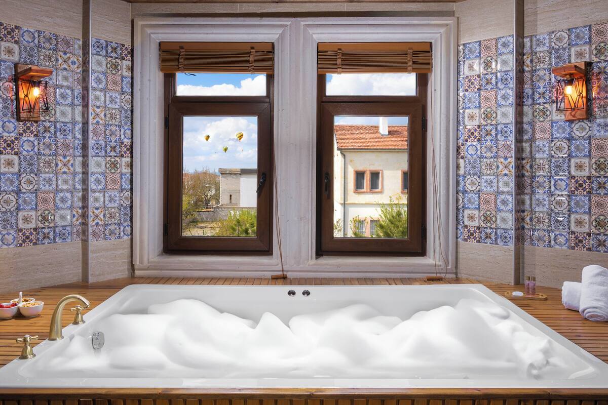 A bathroom with a large bathtub, two windows with blinds, and patterned tile walls.