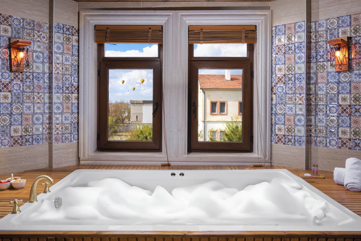 Ein Badezimmer mit einer großen Badewanne, zwei Fenstern mit Jalousien und Wandfliesen mit Muster.