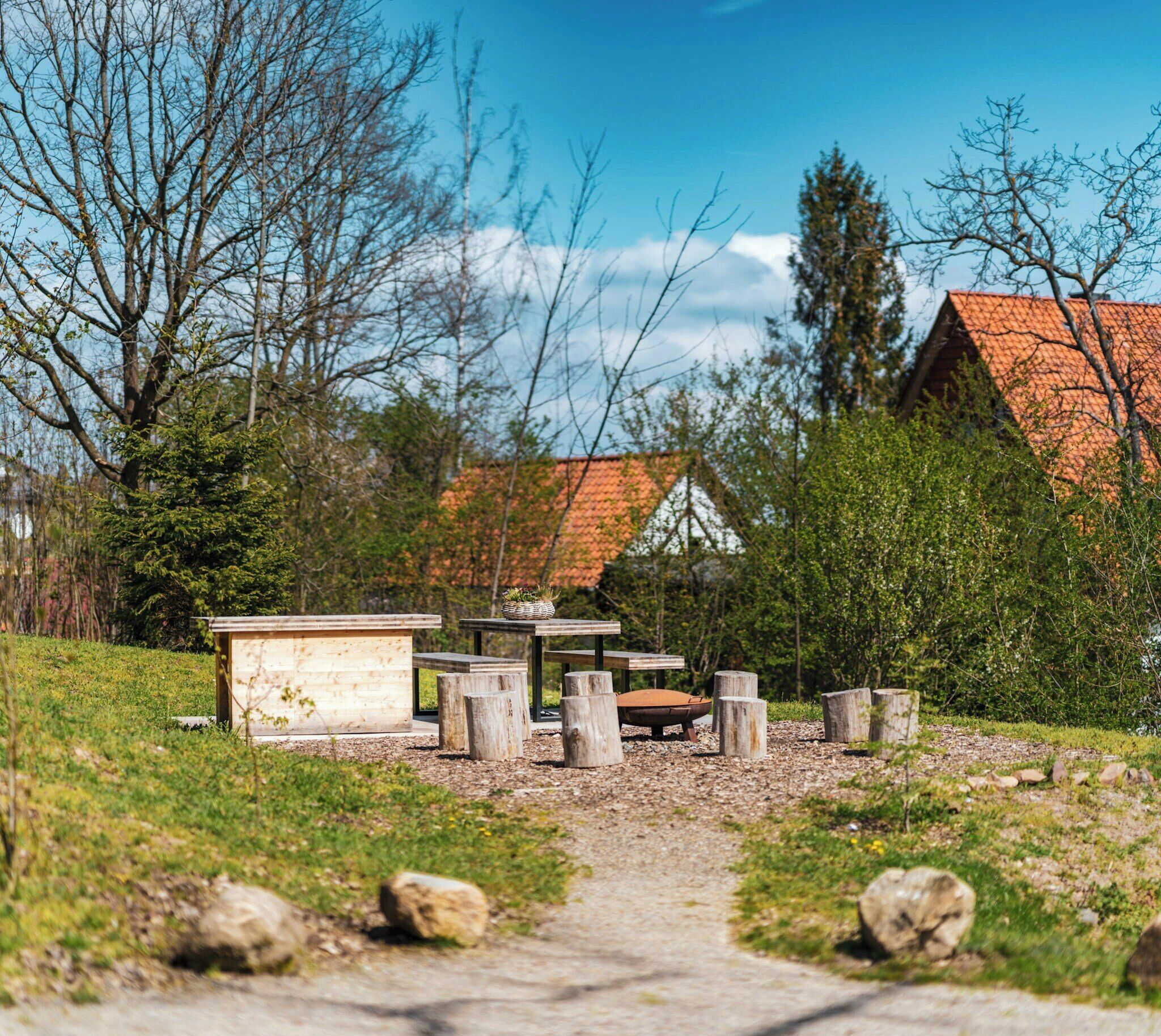 Een rustieke buitenruimte met houten banken, een klein houten gebouw en een pad naar een huis met een rood dak.