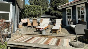Ett trädäck med utemöbler, en planteringslåda i sten och utsikt över ett hus med garage och ett stort träd.