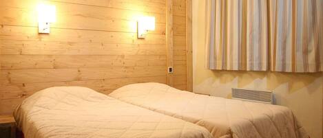 Una habitación con paredes de madera, dos camas con ropa blanca, una cortina a rayas y dos luces fijadas a la pared.
